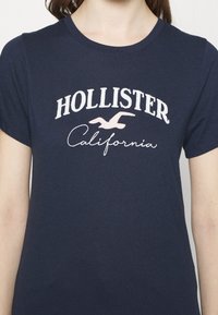 Námořnické bavlněné tričko s nápisem "HOLLISTER" v bílé barvě a "California" v růžovém písmu se siluetou ptáka mezi nimi. Krátké rukávy.