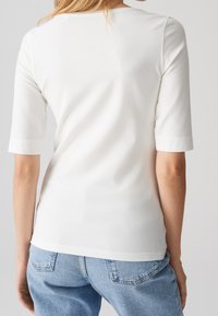 Femme portant un t-shirt blanc uni à manches courtes et un jean bleu clair, vue de dos avec les bras détendus sur les côtés.