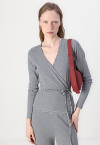 Pruhovaná, dlhá rukávová blúzka v šedej a bielej látke, s výstrihom do V a viazaním v oblasti pása, kombinovaná s červenou ramennou taškou.