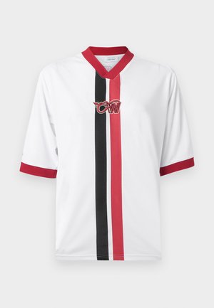 Žena nosiaca veľkú bielu košeľu s červenými a čiernymi pruhmi, modré džínsy, čierne sandále na podpätku, slnečné okuliare a nesie čiernu kabelku na ramene.