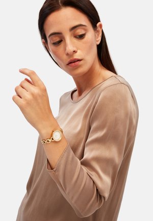 Montre-bracelet en or avec un cadran rond, design de chaîne maillée, et un cadran blanc, portée sur un poignet nu. Un haut à manches longues de couleur neutre est visible.