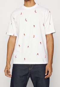 Vit kortärmad t-shirt med utspridda små silhuetter av basketspelare i rött och tryckta bokstaven "J", bärs med mörka jeans.