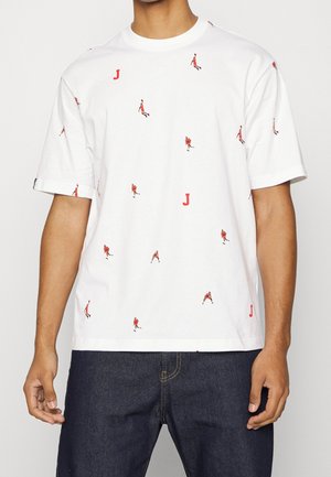 Camiseta blanca de manga corta con siluetas pequeñas de jugadores de baloncesto rojos esparcidas y estampados de la letra "J", combinada con jeans oscuros.