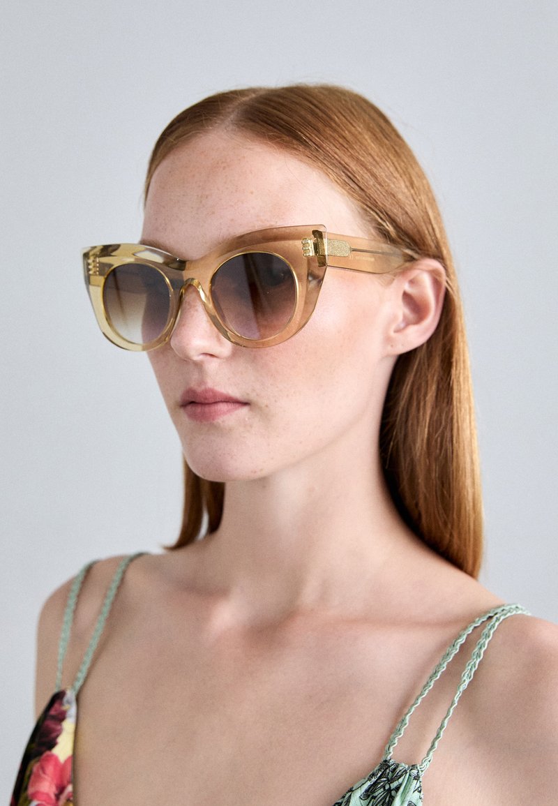Lunettes de soleil en forme d'œil de chat avec des montures transparentes jaunes, des verres dégradés marron, des accents dorés sur les branches et une finition brillante.