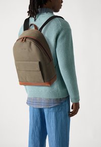 Olivengrønn ryggsekk med en rund form, lærdetaljer og dobbelt glidelås. Båret mot en lys blå genser og løse denimbukser.
