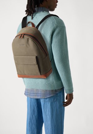 Un sac à dos vert olive à la forme arrondie, avec des accents en cuir et des zips doubles. Porté avec un pull bleu clair et un pantalon en denim ample.