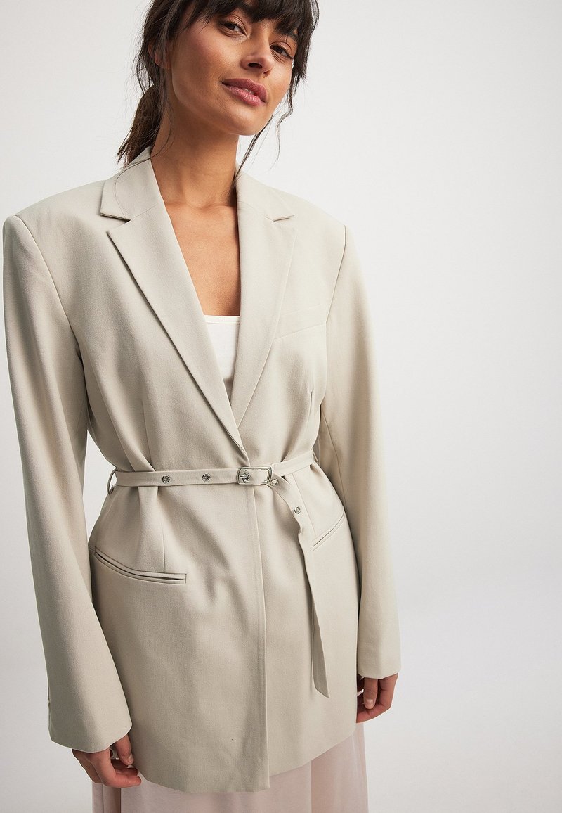 Beige tailored blazer with a belted waist, notch lapels, single breast pocket, and two front pockets, featuring smooth fabric and a structured fit.