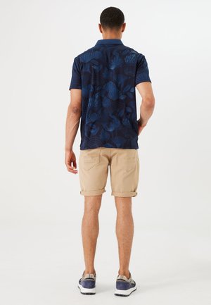 Marineblauw polo shirt met korte mouwen en een bladerenpatroon, gecombineerd met lichtbeige opgerolde shorts. Draag met grijze en blauwe sportschoenen.