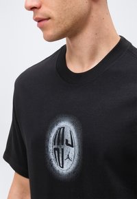 Άνδρας που φοράει μαύρο T-shirt με λευκό οβάλ γραφικό και στυλιζαρισμένα γράμματα και μικρό λογότυπο Jumpman τυπωμένο στο στήθος.
