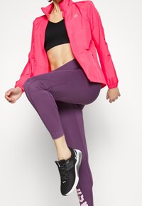 Chaqueta ligera de color rosa brillante con puños elásticos, combinada con leggings morados y zapatillas deportivas negras con acentos blancos.
