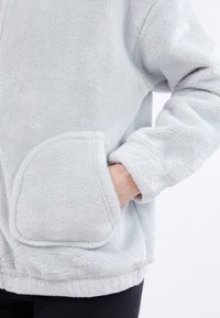 Veste en polaire gris clair, dotée d'une grande poche avant et de manches à côtes. Texture douce avec une coupe décontractée et un ourlet arrondi.