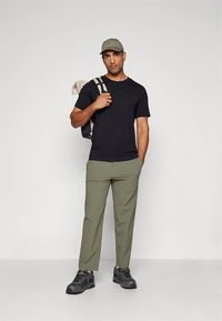 Sort t-shirt, olivengrønne bukser, grå sneakers og en lysegrøn kasket. En beige rygsæk hviler på skulderen med stribede remme.