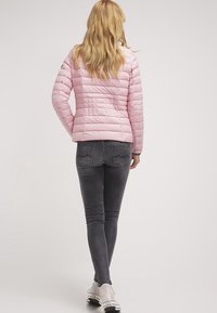 Pastelrosa quilted pufferjakke med en tætsiddende pasform, parret med grå skinny jeans og sølvfarvede sneakers, set fra ryggen.