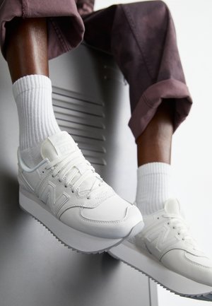 Personne portant des baskets blanches New Balance et des chaussettes blanches côtelées, assise avec les jambes pliées, portant un pantalon foncé retroussé.