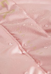 Rosa satin stof har vanddråber med synlige quiltede sektioner. Prægede guldtekst står "Ted Baker" langs sømmen.