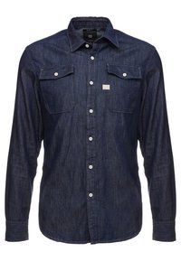 Tmavě modrá džínová košile s dlouhými rukávy, dvěma kapsami na hrudi, zapínáním na knoflíky vpředu a malou nášivkou nad levou kapsou. Hladká textura.