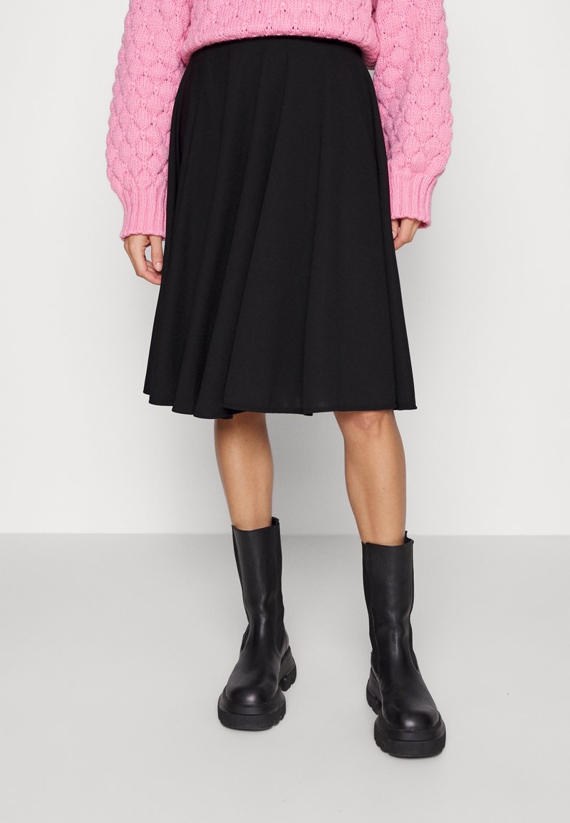 Personne portant une jupe patineuse noire longueur genou, un pull en maille torsadée rose, et des bottes en cuir noires épaisses, debout à l'intérieur.