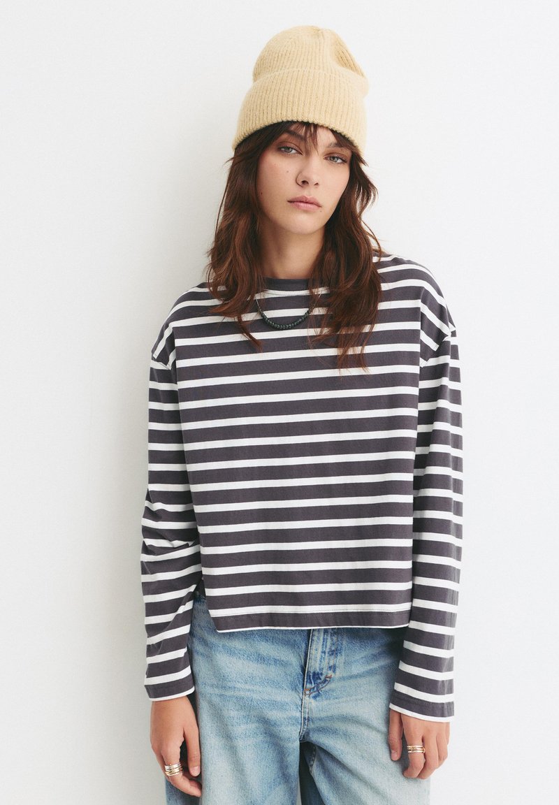 Gestreiftes langärmliges Oberteil aus navyblauem und weißem Baumwollstoff mit einem Bootsausschnitt und überschnittenen Schultern. Kombiniert mit hochgeschnittenen Jeans aus Denim.