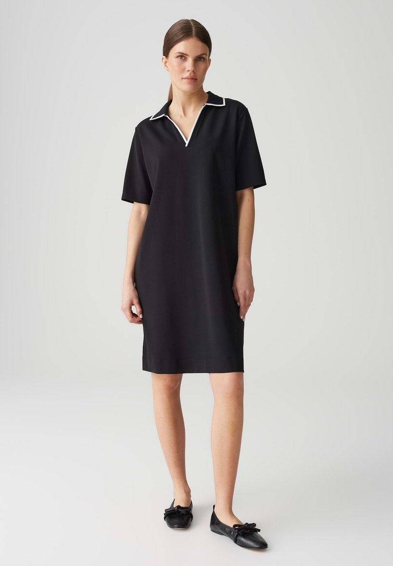 Schwarzes Kleid mit kurzen Ärmeln und einem Polokragen, mit weißem Besatz, glatter Textur und geradem Schnitt. Getragen mit schwarzen Ballerinas.