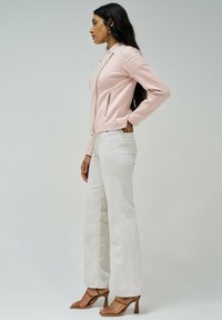 Svetlo roza usnjena jakna z diagonalnimi zadrgami in teksturiranimi ramenimi poudarki, v kombinaciji z off-white širokimi hlačami in rjavimi sandali s peto.