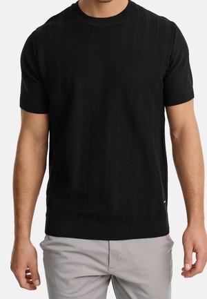 Mężczyzna ubrany w czarny, krótkim rękawem, o fakturze sweter z dzianiny oraz jasnoszare spodnie, stojący na jednolitym tle.