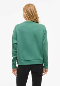 Femme aux longs cheveux blonds portant un sweat-shirt vert à manches longues et un jean noir, se tenant de dos contre un fond blanc.