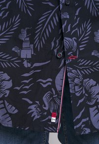 Chemise noire à boutons avec motifs de feuilles tropicales violettes et de fleurs, bordure rouge et blanche à l'intérieur de la patte de boutonnage, portée avec un jean bleu.