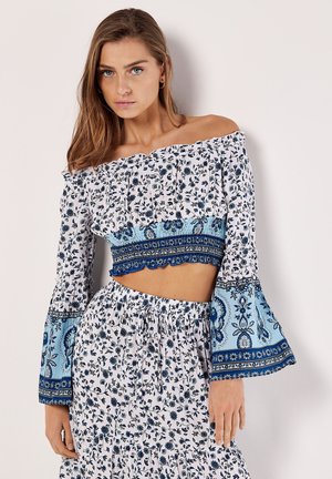 Besluttet cropped top med off-the-shoulder-design og blomsterprint i hvid og blå. Har flæser på ærmerne og en smocked bodice, kombineret med en matchende nederdel.