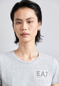Tricou gri din bumbac, cu guler rotund și mâneci scurte. Prezintă un logo EA7 texturat pe piept, cu țesătură netedă și un model discret de heather.