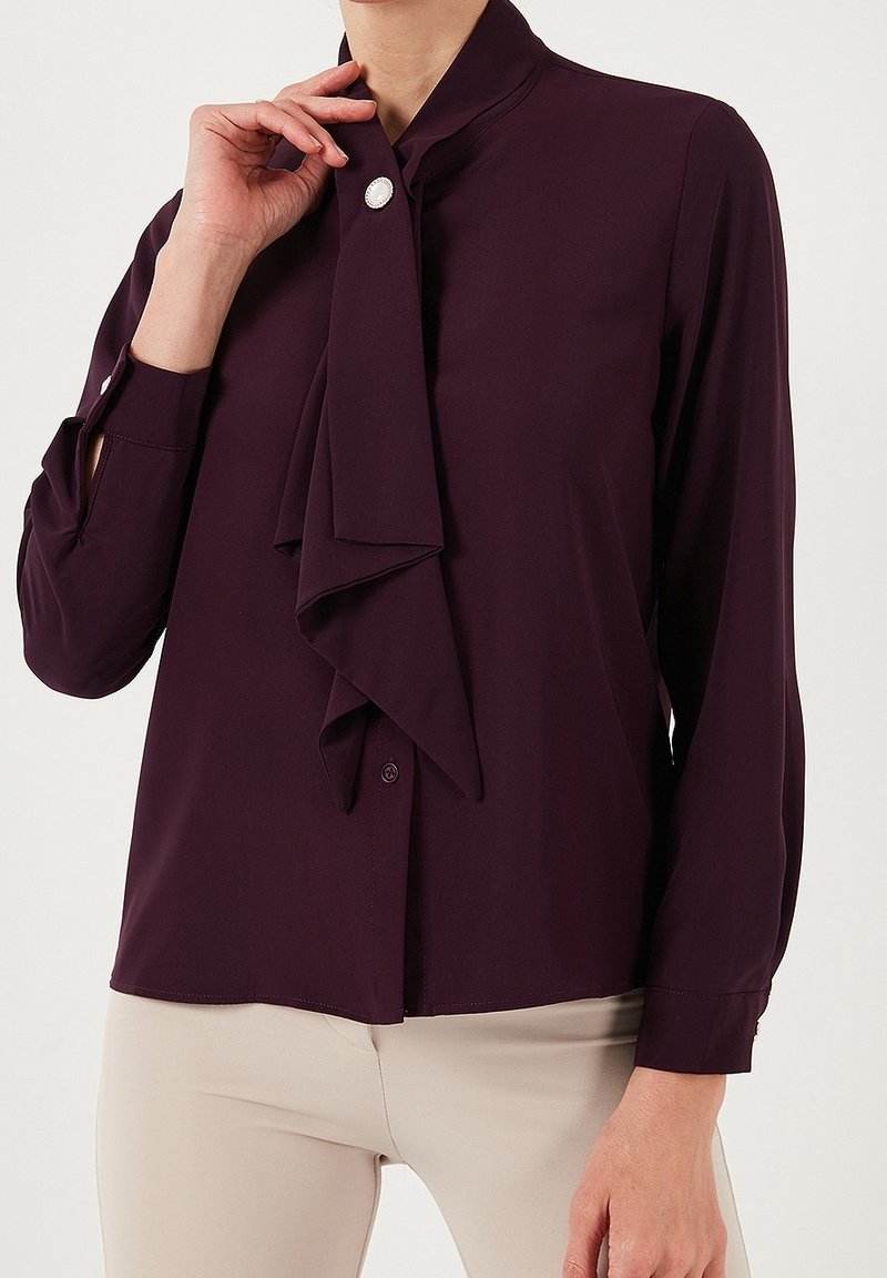 Femme portant un chemisier violet foncé avec une cravate et un bouton en perle, associé à un pantalon beige, posant avec une main près du col.
