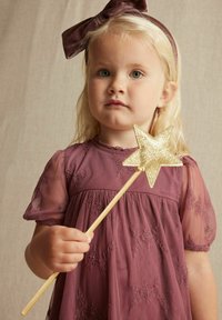 Ein Kind in einem lavendelfarbenen Kleid mit durchsichtigen Puffärmeln hält einen goldenen Sternenstab, der mit Glitzereffekten verziert ist. Schlichter Hintergrund in neutralen Tönen.