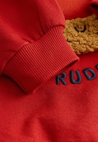 Czerwona bluza z krótkim rękawem z teksturowanym motywem brązowego niedźwiedzia z przodu, z ribbowymi mankietami i haftem "RUDY" w granatowym kolorze. Miękka tkanina.