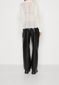 Vit spetsblus med långa ärmar och rund halsringning, kombinerad med högmidjade svarta läderbyxor och svarta skor. Texturerade detaljer synliga.