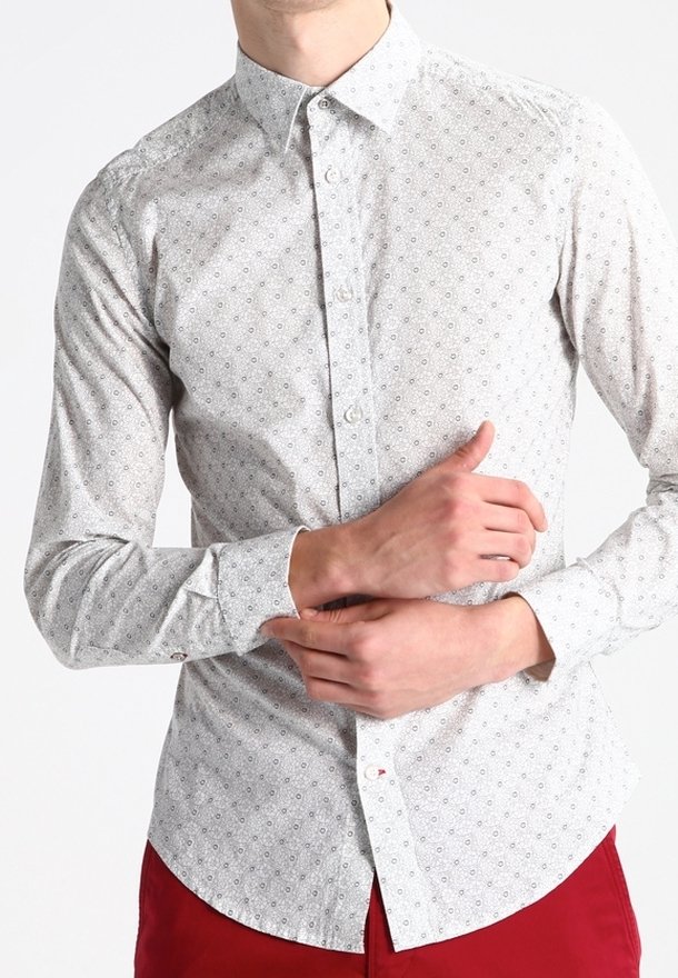 Homme portant une chemise gris clair à boutons avec de petits motifs circulaires et un pantalon rouge, ajustant le poignet gauche de la chemise avec les deux mains.