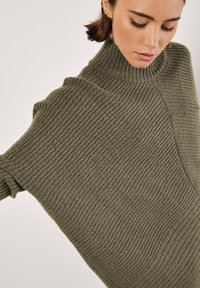 Sweter z dzianiny z wysokim kołnierzem w kolorze oliwkowym, z prążkowaną teksturą, w oversize'owym kroju i szerokimi rękawami. Szczegóły szwu dodają wymiaru.
