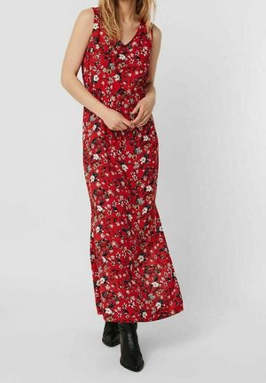 Kobieta ubrana w czerwoną, długą sukienkę bez rękawów z kwiatowym wzorem, w czarnych botkach za kostkę, stojąca na jasnym, jednolitym tle.