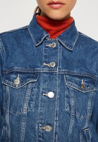 Denim jakna s klasičnim krojem, plave boje, srebrnim dugmadi, dva džepa na grudima i blago iznošenom teksturom. Crveni rolak ispod.