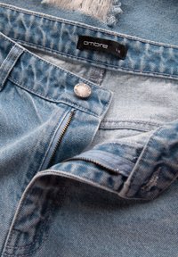 Jeans de mezclilla azul claro con una cremallera parcialmente abierta, botón metálico y etiqueta negra que muestra la marca "ombre" y talla L 33.