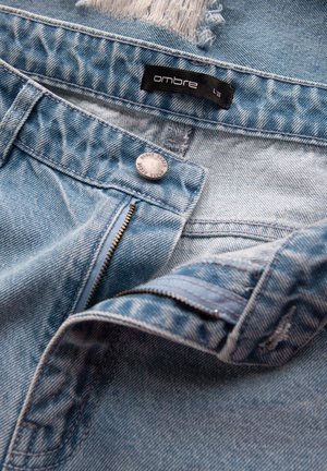 Jean en denim bleu clair avec fermeture éclair, bouton argenté et détails déchirés. L'étiquette indique "ombre" taille L 33. Tissu texturé.