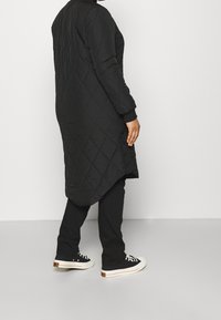 Person som bär en lång svart vadderad kappa, svarta byxor och svarta låga sneakers med vita sulor, som står mot en enfärgad bakgrund.