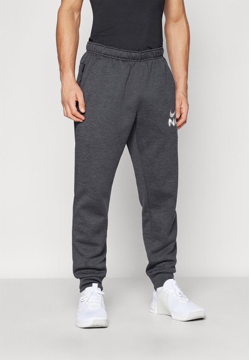 Grå fleece-joggers med elastisk midjeband, utrustade med sidofickor och en vit logotyp på vänster lår. Bärs med vita sportskor.