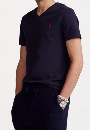Homme portant un t-shirt à col en V bleu marine avec un logo rouge, un pantalon bleu marine avec cordon de serrage, et une montre-bracelet accompagnée de bracelets tressés blancs au poignet gauche.