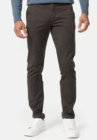 Tummanruskeat chinos-housut, jotka on valmistettu sileästä kankaasta, joissa on vartalonmyötäinen leikkaus ja etutaskut, yhdistettynä valkoisiin tennareihin.