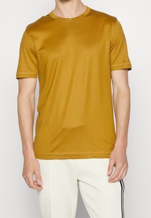 Homme portant un t-shirt jaune moutarde uni à manches courtes et un pantalon blanc avec une bande noire latérale sur un fond uni.