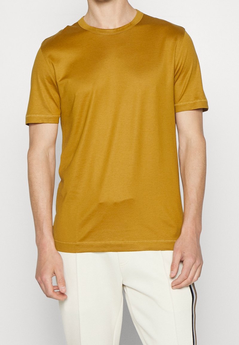 Homme portant un t-shirt jaune moutarde uni à manches courtes et un pantalon blanc avec une bande noire latérale sur un fond uni.