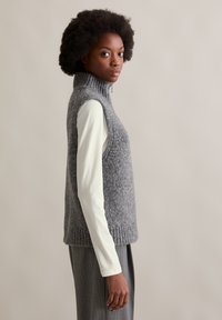 Frau mit Afro-Frisur trägt graue ärmellose Strickweste über weißem Langarmshirt und grauer Nadelstreifenhose, seitlich stehend.