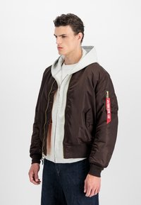 Hnedá saténová bomber bunda s rebrovanými manžetami a spodným lemom, pod ktorou je šedá mikina s kapucňou, vrecko na rukáve a zlatý zips.