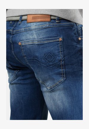 Jeans in denim blu scuro con un leggero sbiadimento. Presentano un design ricamato sulla tasca, una etichetta in pelle marrone sulla vita e rivetti di colore rame.