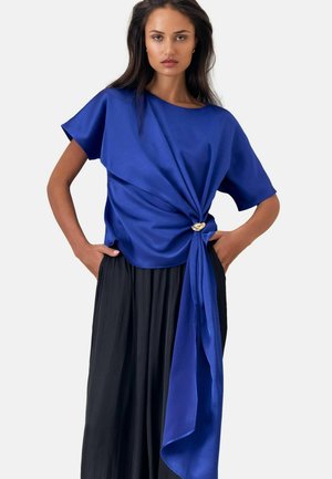 Frau mit langen dunklen Haaren trägt ein blaues Satin-Kurzarmtop mit seitlichem Knoten und einen schwarzen plissierten Rock, posiert mit Händen in den Taschen.