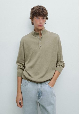 Olive green pullover with a high collar and three-button placket, made of soft fabric. Paired with light blue, relaxed-fit jeans.
